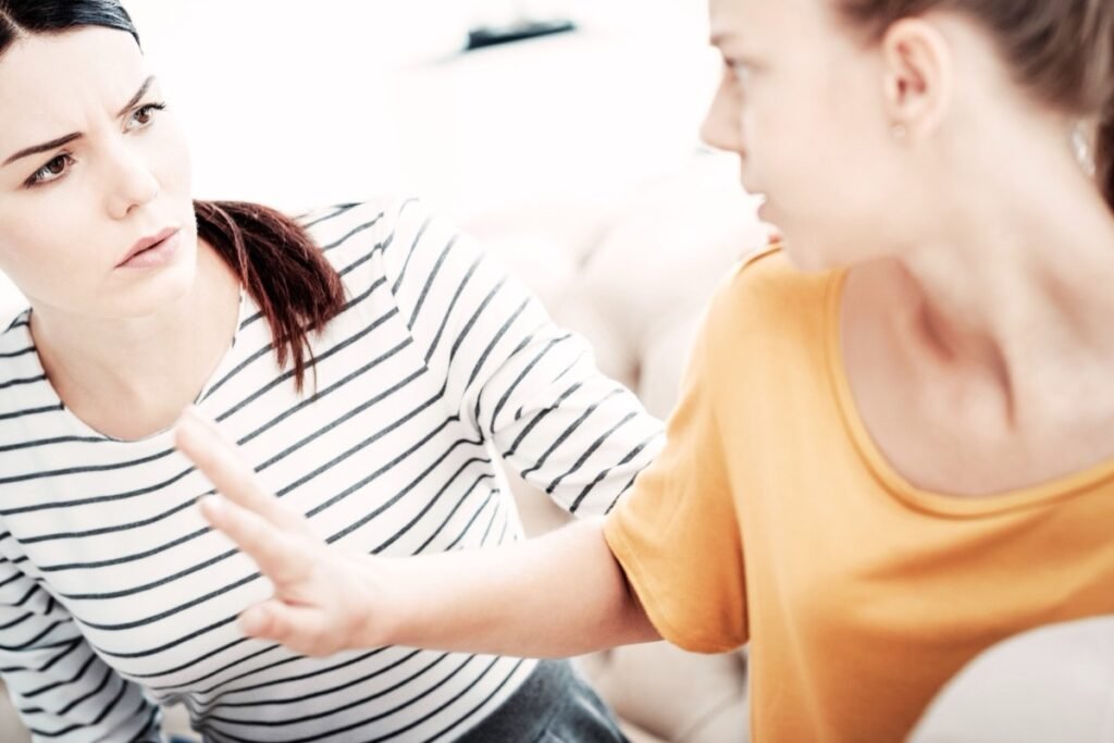 Female in black and white stripe top who is worried is speaking to a woman in an orange top with blonde hair who is being defensive
