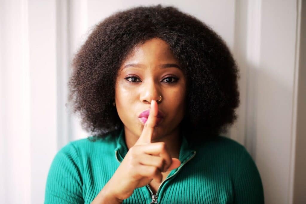 woman with dark brown hair and green jumper making a shh motion with her finger over her lips